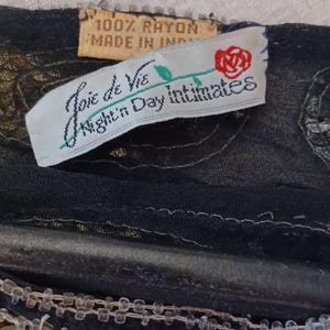 Puede incluir: Primer plano de una prenda negra con lentejuelas doradas y plateadas. La prenda tiene una etiqueta que dice "Joie de Vie Night Day Intimates" y "100% Rayon Made in India". El fondo es una tela blanca texturizada.