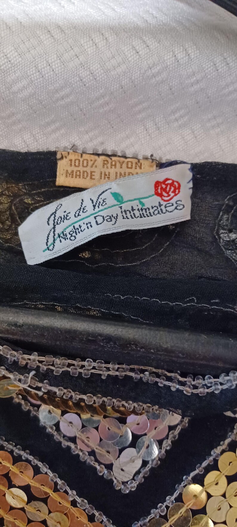 Puede incluir: Primer plano de una prenda negra con lentejuelas doradas y plateadas. La prenda tiene una etiqueta que dice "Joie de Vie Night Day Intimates" y "100% Rayon Made in India". El fondo es una tela blanca texturizada.