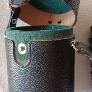 Puede incluir: Una funda de c&aacute;mara de cuero negro con un interior de terciopelo verde y una correa. La funda est&aacute; abierta y la tapa est&aacute; volteada hacia atr&aacute;s.