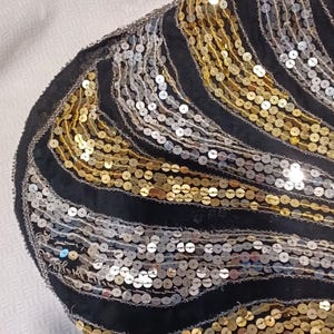 Puede incluir: Tela negra con lentejuelas doradas y plateadas en un patr&oacute;n ondulado. Las lentejuelas est&aacute;n cosidas sobre una base de tela negra. El dise&ntilde;o recuerda al estilo Art D&eacute;co.