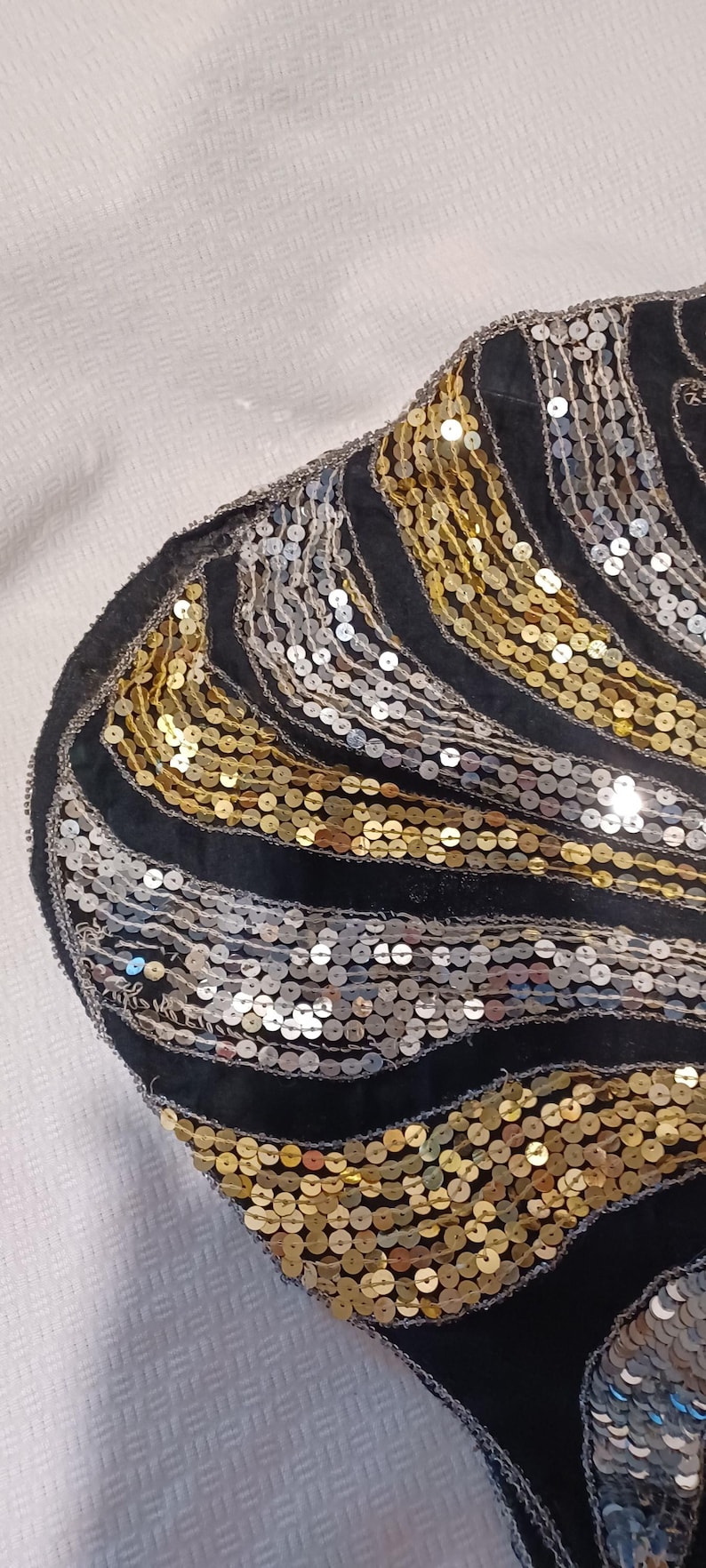 Puede incluir: Tela negra con lentejuelas doradas y plateadas en un patr&oacute;n ondulado. Las lentejuelas est&aacute;n cosidas sobre una base de tela negra. El dise&ntilde;o recuerda al estilo Art D&eacute;co.