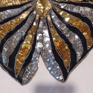 Puede incluir: Primer plano de una prenda negra con lentejuelas con un dise&ntilde;o de mariposa. El dise&ntilde;o presenta lentejuelas doradas y plateadas dispuestas en un patr&oacute;n de rayas, con una gran lentejuela dorada en el centro.