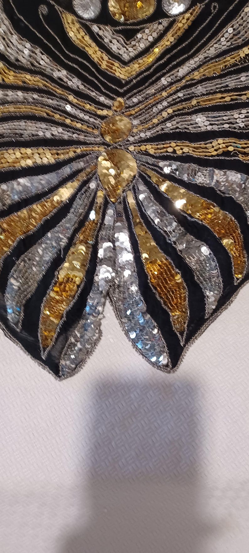 Puede incluir: Primer plano de una prenda negra con lentejuelas con un dise&ntilde;o de mariposa. El dise&ntilde;o presenta lentejuelas doradas y plateadas dispuestas en un patr&oacute;n de rayas, con una gran lentejuela dorada en el centro.