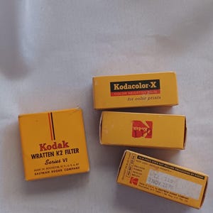 May include: Four vintage Kodak film boxes in yellow. The boxes include "Kodacolor-X" and "Kodak Wrattem K2 Filter" labels. The boxes are on a white surface. The boxes are from Eastman Kodak Company.