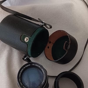 Puede incluir: Un objetivo de c&aacute;mara negro y plateado con el texto "1:2.8/135 Asahi Opt. Co. Lens Made in Japan 1318015 Super-Takumar" impreso en el lateral. El objetivo est&aacute; en una funda de cuero negro con forro verde.