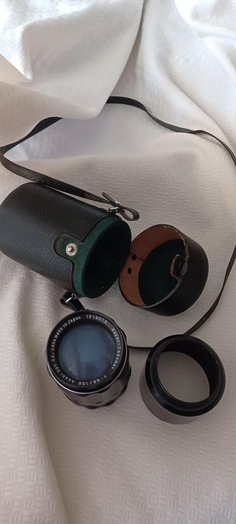 Puede incluir: Un objetivo de c&aacute;mara negro y plateado con el texto "1:2.8/135 Asahi Opt. Co. Lens Made in Japan 1318015 Super-Takumar" impreso en el lateral. El objetivo est&aacute; en una funda de cuero negro con forro verde.