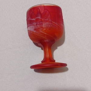 May include: A red glass cordial glass with an eagle design. The glass has a stem and a round base. The glass is made of red glass with white marbling. The glass is likely vintage.