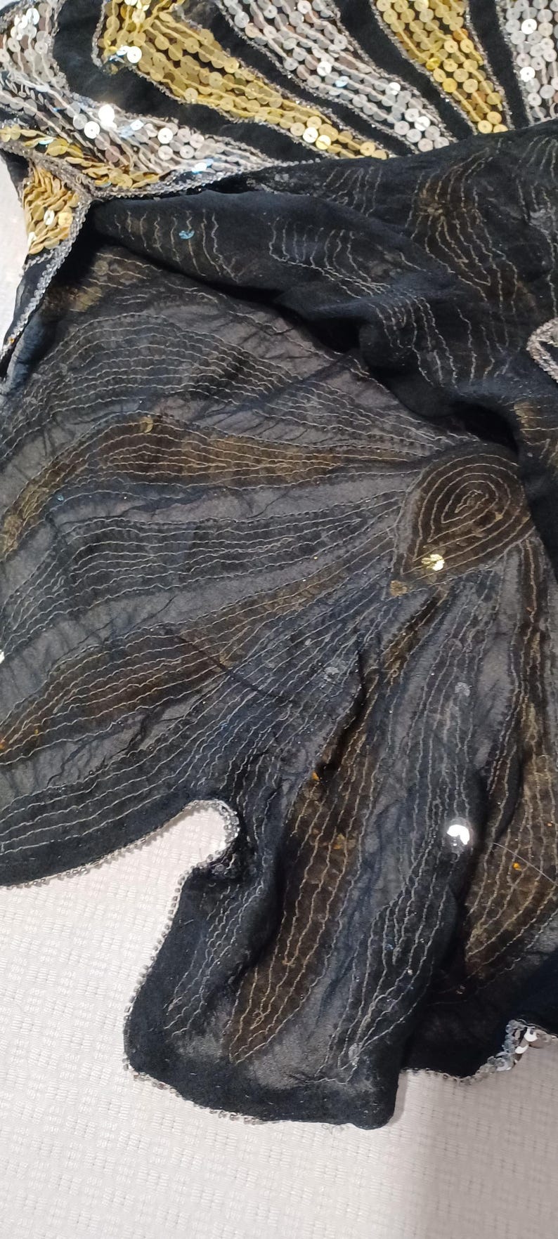 Puede incluir: Tela negra con un patr&oacute;n de l&iacute;neas curvas y lentejuelas. El borde superior tiene un dise&ntilde;o de lentejuelas doradas y plateadas en un patr&oacute;n de rayas. La tela est&aacute; drapeada sobre una superficie blanca.