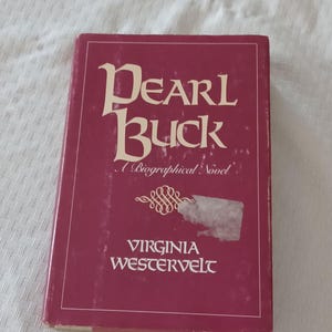 May include: A maroon book titled "Pearl Buck" with the subtitle "A Biographical Novel" by Virginia Westervelt. The book has a white sticker on the cover and is set against a white patterned background.