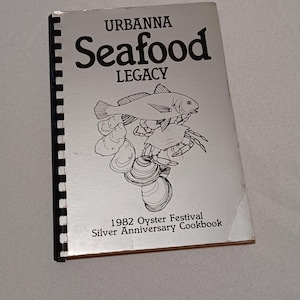 May include: A silver-colored cookbook titled "Urbanna Seafood Legacy" with a spiral binding. The cover features illustrations of fish, shrimp, and oysters. Text on the cover reads "1982 Oyster Festival Silver Anniversary Cookbook."