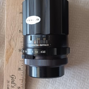 Puede incluir: Un objetivo de c&aacute;mara negro con una banda plateada y una etiqueta blanca que dice "PASSD". El objetivo tiene una escala de profundidad de campo con n&uacute;meros del 22 al 16, 8, 5.6, 43.5, y una escala de distancia con n&uacute;meros del 22161156 al 56111622. El objetivo tambi&eacute;n tiene un anillo de enfoque con n&uacute;meros del 100 al 50, 30 y 15.