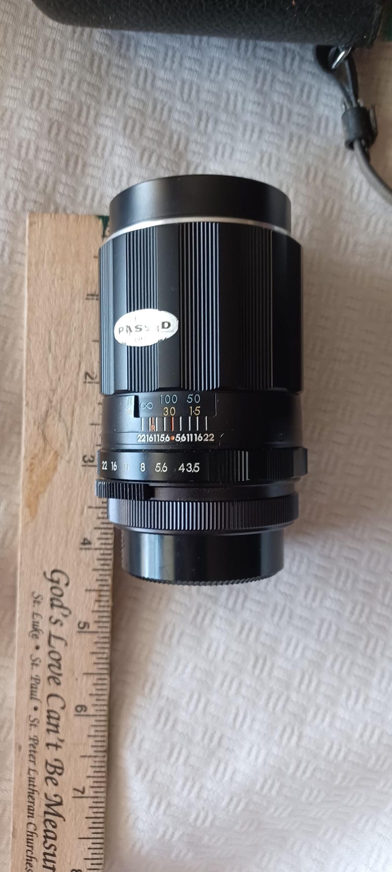 Puede incluir: Un objetivo de c&aacute;mara negro con una banda plateada y una etiqueta blanca que dice "PASSD". El objetivo tiene una escala de profundidad de campo con n&uacute;meros del 22 al 16, 8, 5.6, 43.5, y una escala de distancia con n&uacute;meros del 22161156 al 56111622. El objetivo tambi&eacute;n tiene un anillo de enfoque con n&uacute;meros del 100 al 50, 30 y 15.