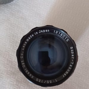 Puede incluir: Un objetivo de c&aacute;mara negro con el texto "Super-Takumar 1:3.5/135 Asahi Opt. Co., Lens made in Japan 1318015" impreso alrededor del borde.