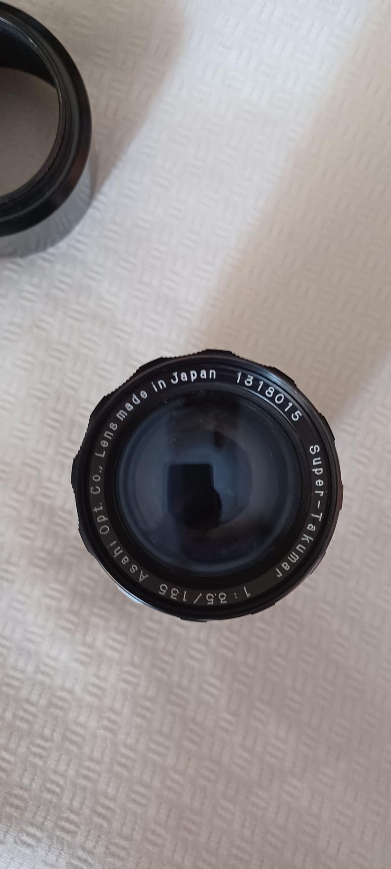 Puede incluir: Un objetivo de c&aacute;mara negro con el texto "Super-Takumar 1:3.5/135 Asahi Opt. Co., Lens made in Japan 1318015" impreso alrededor del borde.