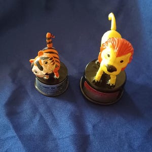 May include: Two vintage toy figurines of jungle animals. One is a tiger with orange and black stripes, and the other is a lion with a yellow body and red mane. Both are on black bases with text.