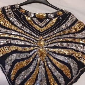 Puede incluir: Un top negro con lentejuelas doradas y plateadas, colgado de una percha negra. El dise&ntilde;o presenta l&iacute;neas radiantes de lentejuelas, creando un patr&oacute;n de rayos de sol. El top tiene un escote redondo y un adorno central.