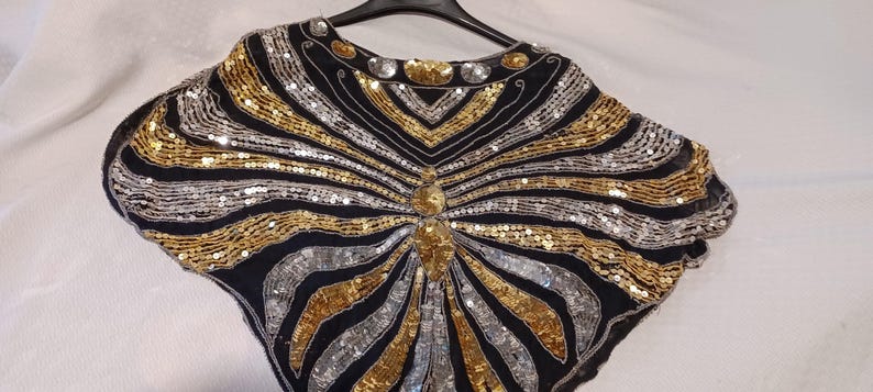 Puede incluir: Un top negro con lentejuelas doradas y plateadas, colgado de una percha negra. El dise&ntilde;o presenta l&iacute;neas radiantes de lentejuelas, creando un patr&oacute;n de rayos de sol. El top tiene un escote redondo y un adorno central.