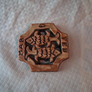 May include: A brown ceramic plaque featuring a relief design of two fish swimming in opposite directions. The text MAR is embossed on the left side and FEB on the right side. A small elephant icon is centered at the top of the octagonal frame.