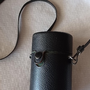 Puede incluir: Una funda de c&aacute;mara de cuero negro con una correa y un cierre de metal. La funda tiene una superficie texturizada.