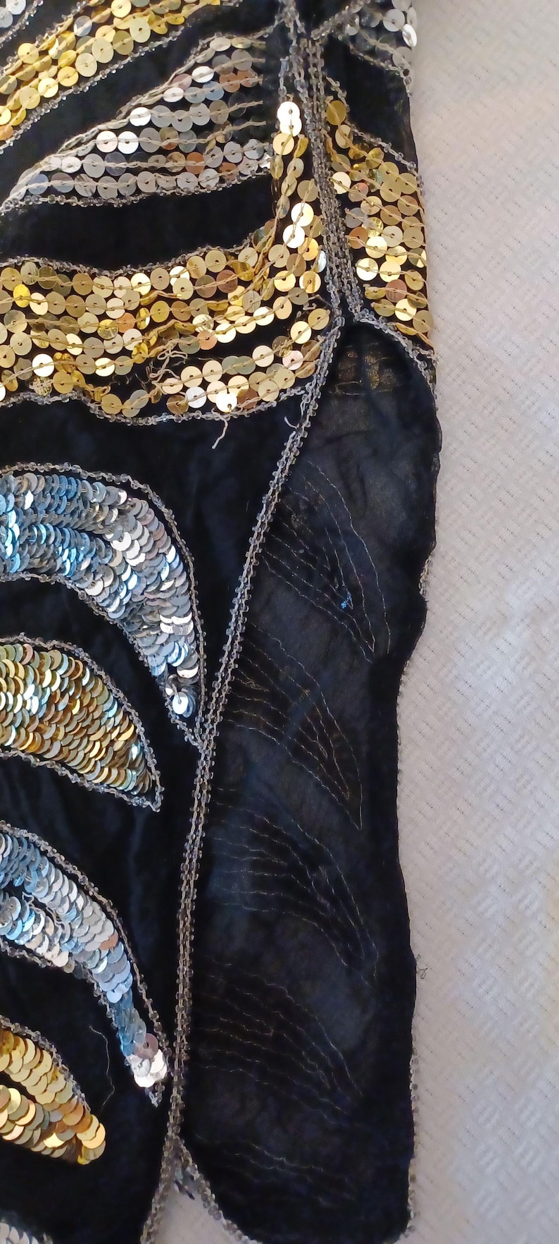 Puede incluir: Tela negra adornada con lentejuelas doradas y plateadas. Las lentejuelas est&aacute;n dispuestas en un patr&oacute;n similar a una hoja, con lentejuelas plateadas a la izquierda y doradas a la derecha. Los bordes est&aacute;n ribeteados con hilo plateado.