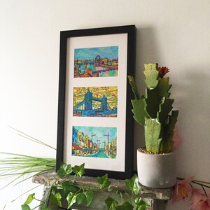 May include: A black framed print with three colorful images of London landmarks. The top image shows the London Eye, the middle image shows Tower Bridge, and the bottom image shows a street scene.