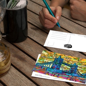 May include: A colorful postcard featuring a stylized illustration of Tower Bridge in London, England. The postcard is on a wooden table with a cup of coffee and a pen.