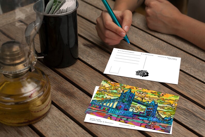 May include: A colorful postcard featuring a stylized illustration of Tower Bridge in London, England. The postcard is on a wooden table with a cup of coffee and a pen.