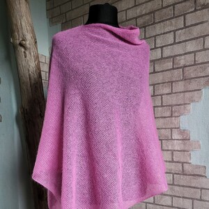May include: A light pink knitted poncho displayed on a mannequin. The poncho has a cowl neck and a loose, flowing design. The background features a brick wall and a wooden log.