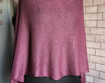 Op de afbeelding: Een pruimkleurige poncho met een col wordt getoond op een zwarte mannequin. De gebreide stof drapeert over de schouders en heeft een gelaagd ontwerp. De achtergrond is voorzien van een bakstenen muur en een houten paal.