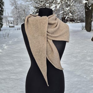 Może przedstawiać: Beżowy szalik jest narzucony na czarny manekin. Szalik ma trójkątny kształt z długimi, spiczastymi końcami. Tło to zaśnieżona sceneria na zewnątrz z drzewami i ścieżką.
