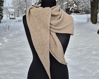 May include: A beige scarf is draped over a black mannequin. The scarf has a triangular shape with long, pointed ends. The background is a snowy outdoor scene with trees and a pathway.