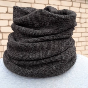May include: A dark grey knitted cowl scarf. The scarf is made of a thick, textured yarn and is designed to be worn around the neck. The cowl is shown against a brick wall background.