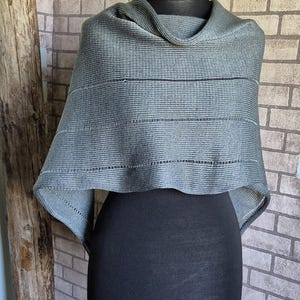 Könnte beinhalten: Ein grauer Strickschal mit einem Rollkragen, der auf einer schwarzen Schaufensterpuppe präsentiert wird. Der Schal hat horizontale Streifendetails. Der Hintergrund zeigt eine Backsteinmauer und einen Holzpfosten.