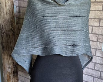 May include: A gray knitted shawl with a cowl neck, displayed on a black mannequin. The shawl has horizontal striped detailing. The background features a brick wall and a wooden post.