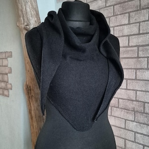 May include: A black knitted hooded scarf on a mannequin. The scarf is triangular and drapes over the shoulders. The background features a brick wall and a wooden accent.