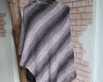 May include: A gray and black striped poncho displayed on a mannequin. The poncho is made of a soft, knitted material and features a diagonal stripe pattern. The mannequin is on a white stand, set against a brick wall.