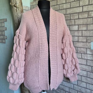 May include: A light pink open-front cardigan featuring a textured knit body and voluminous sleeves adorned with a repeating bobble stitch pattern. The garment has a ribbed collar and ribbed cuffs, displaying a consistent pastel pink yarn construction.