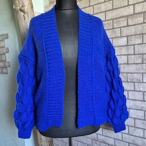 May include: A vibrant royal blue open-front cardigan featuring a chunky knit texture. The garment displays long sleeves adorned with a repeating pattern of raised bobble stitches, a ribbed collar, and ribbed cuffs, presented on a black mannequin.