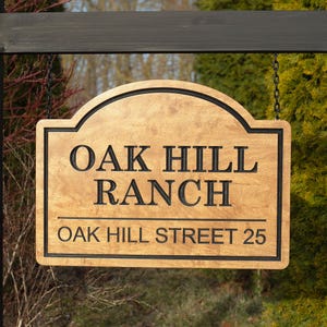 Könnte beinhalten: Holzschild für Oak Hill Ranch mit schwarzer Schrift und Umrandung. Das Schild lautet "OAK HILL RANCH" über "OAK HILL STREET 25". Das Schild hängt an einem Holzpfosten.