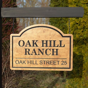May include: Wooden sign for Oak Hill Ranch, with black lettering and border. The sign reads "OAK HILL RANCH" above "OAK HILL STREET 25". The sign is hanging from a wooden post.