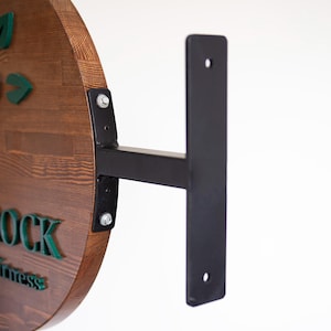 Könnte beinhalten: Ein rundes Holzschild mit grünen Buchstaben und Blattakzenten, montiert an einer schwarzen Metallhalterung. Das Schild hat eine dunkle Holzmaserung und ist mit Schrauben an der Halterung befestigt. Die Halterung ist an einer weißen Wand befestigt.