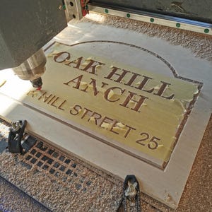 May include: A wooden sign being carved by a CNC machine. The sign reads "OAK HILL ANCH HILL STREET 25" in a serif font. The wood is light-colored, and the machine is actively cutting the letters.