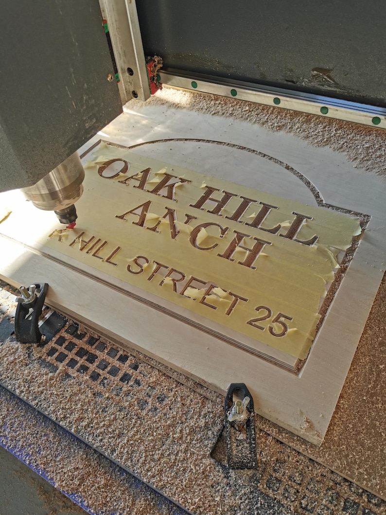 May include: A wooden sign being carved by a CNC machine. The sign reads "OAK HILL ANCH HILL STREET 25" in a serif font. The wood is light-colored, and the machine is actively cutting the letters.
