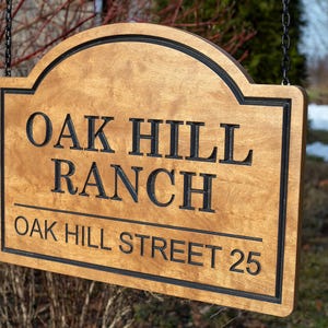 May include: A wooden sign with the words "OAK HILL RANCH" and "OAK HILL STREET 25" in black lettering. The sign has a light brown finish with a black border and is hanging from a chain.
