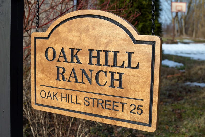 May include: A wooden sign with the words "OAK HILL RANCH" and "OAK HILL STREET 25" in black lettering. The sign has a light brown finish with a black border and is hanging from a chain.