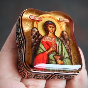 May include: A small, ornate, copper-colored box with a gold-toned image of an angel with wings. The angel is wearing a red and green robe and holding a cup. The box has Cyrillic text around the edges.