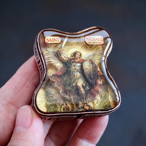 May include: A small, rectangular, copper-toned metal box with a domed, glossy resin top featuring an illustration of Saint Michael the Archangel holding a sword and shield. The text SAINT MICHAEL THE ARCHANGEL is printed on the top surface.