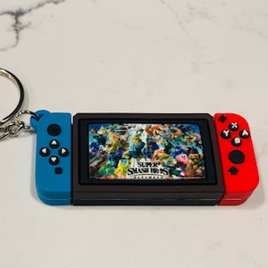 May include: A blue and red Nintendo Switch keychain with a black screen displaying the Super Smash Bros. Ultimate logo and characters. The keychain has a silver key ring.