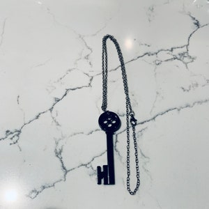 May include: A black key-shaped pendant necklace with a silver chain. The key has a circular design with multiple holes in the center.
