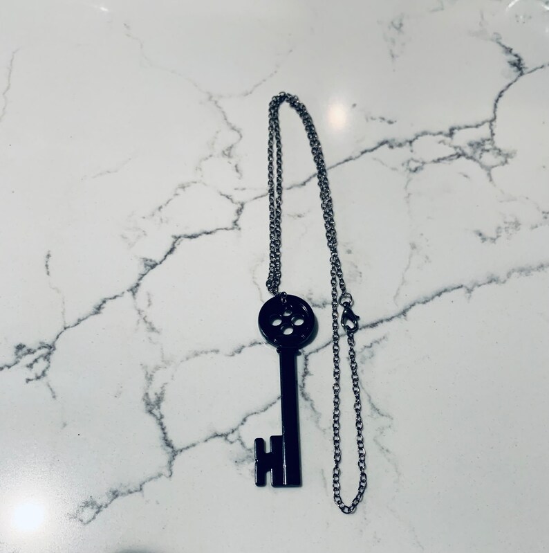 May include: A black key-shaped pendant necklace with a silver chain. The key has a circular design with multiple holes in the center.